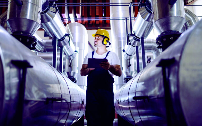 Focused plant worker in overalls, with protective helmet on head and antiphons on ears using tablet for checking machine.