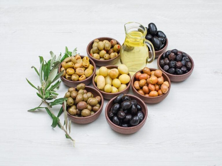 Assorted variety of olives in a clay bowls with olive leaves and a jar of olive oil high angle view on a white wooden background