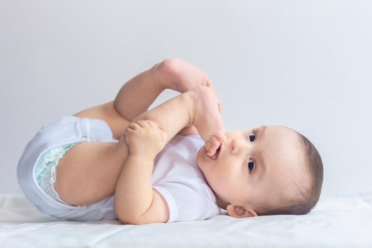 Little hungry baby boy taking feet in mouth. 6-month baby having fun in white bedding. Cute baby lying on bed. Family, new life, childhood concept.